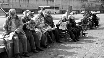 Un gruppo di aziani legge e commenta il giornale in una piazza pubblica.
